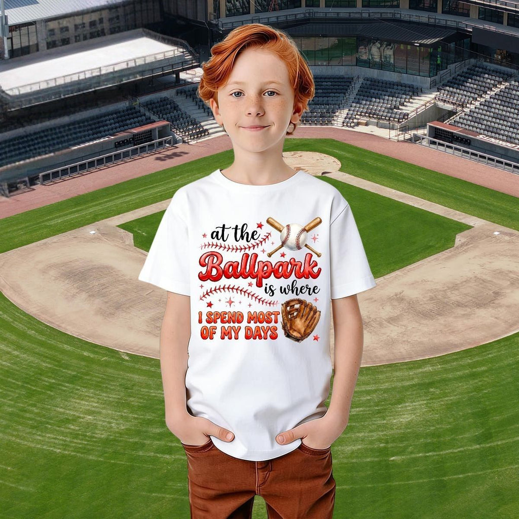 Child wearing a white t-shirt with a baseball-themed design on a baseball field background.