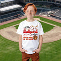 Child wearing a white t-shirt with a baseball-themed design on a baseball field background.