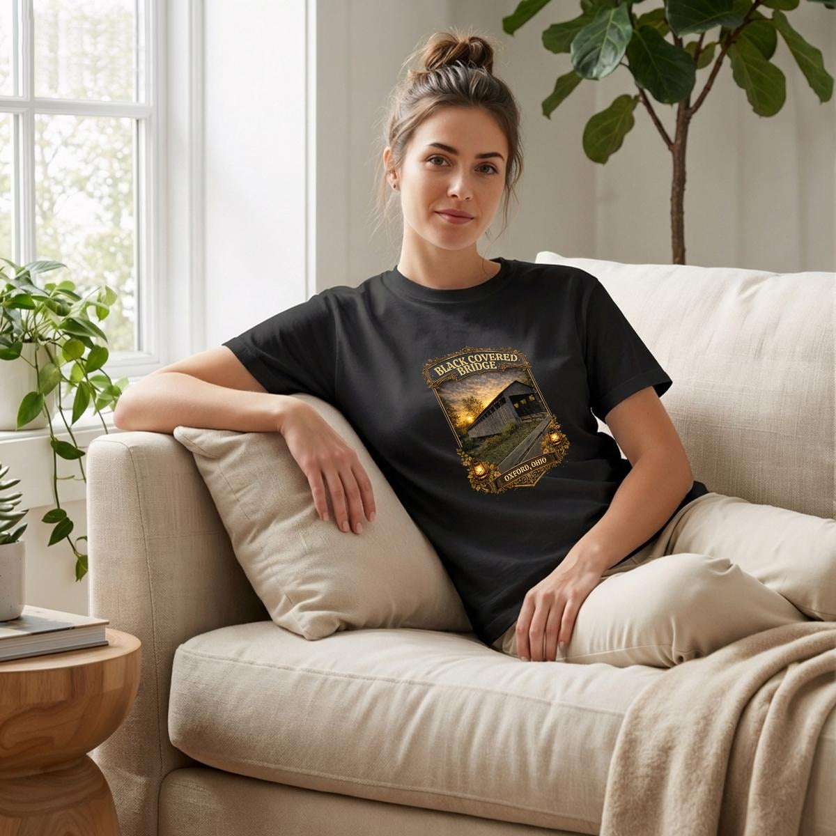 Woman sitting on a couch wearing a black t-shirt with graphic of Black Covered Bridge shirt.