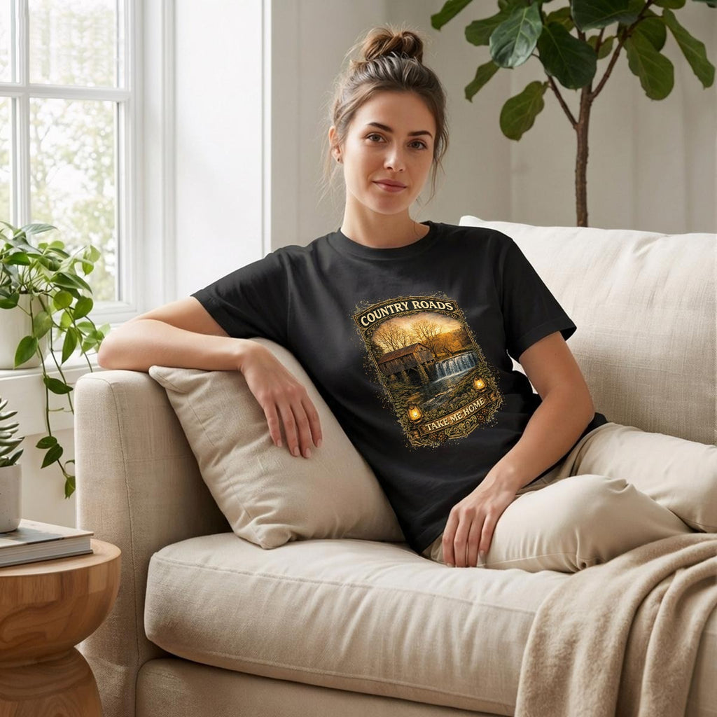Woman sitting on a couch wearing a black t-shirt with graphic of Country Roads-Take Me Home Shirt.