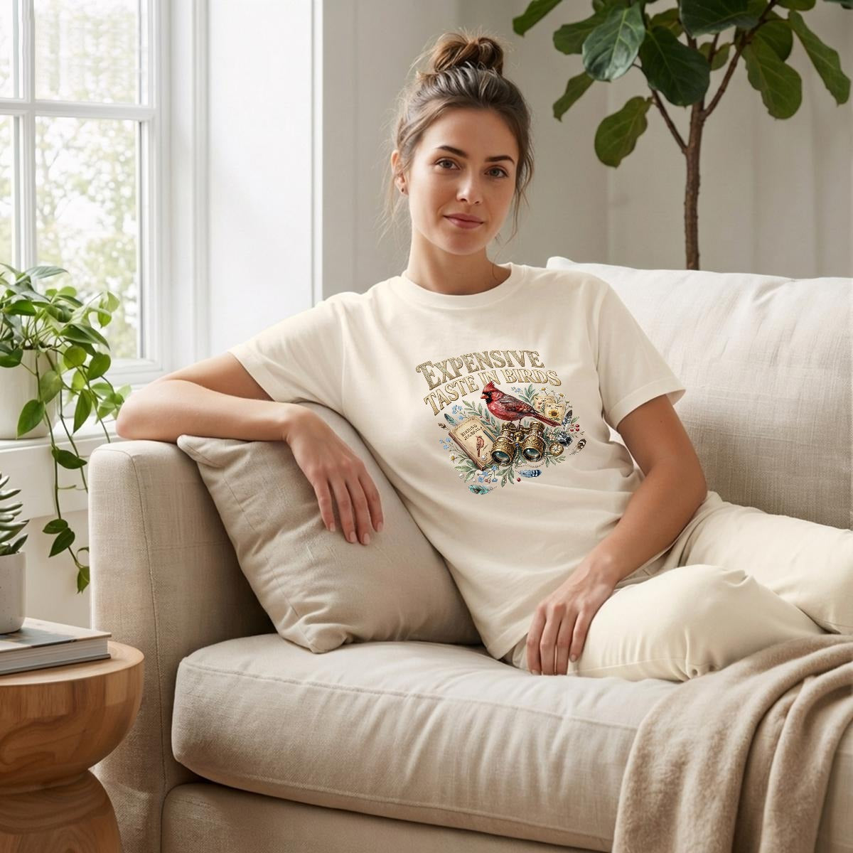 Woman sitting on a couch wearing a natural t-shirt with text about expensive taste in birds, in a bright room.