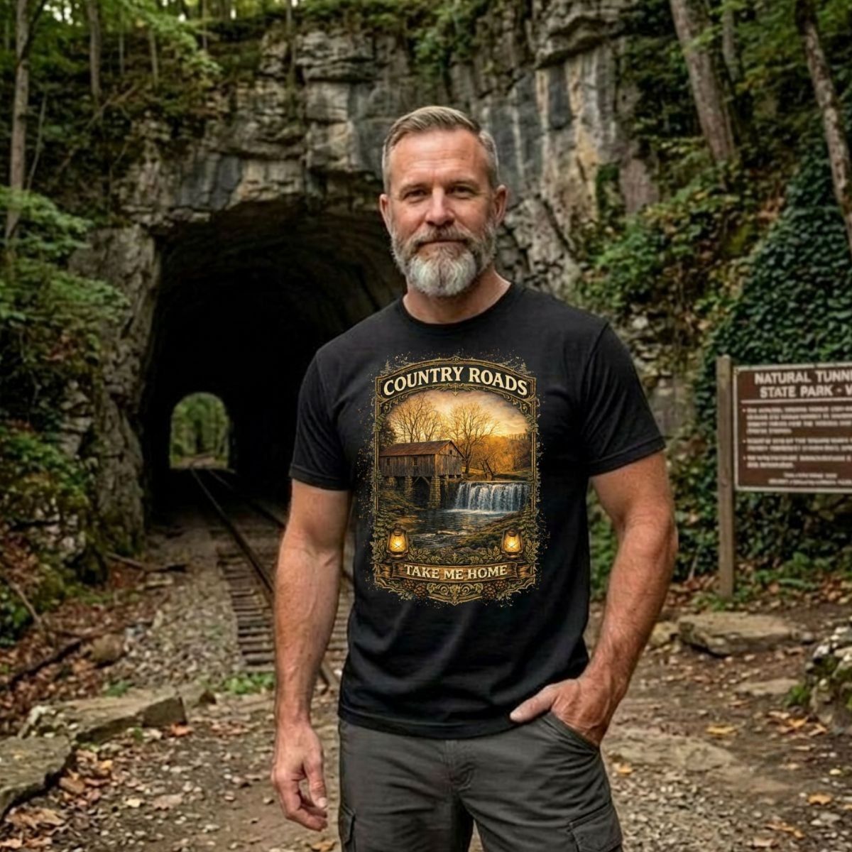 Man wearing a black t-shirt with graphic design - Country Roads-Take Me Home Shirt, in a nature park.
