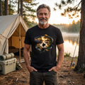 Man wearing a black t-shirt with a nature-themed design, standing in front of a tent and lake.