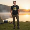 Man standing by a lake with a tent and fire pit, wearing a black t-shirt with a nature design.